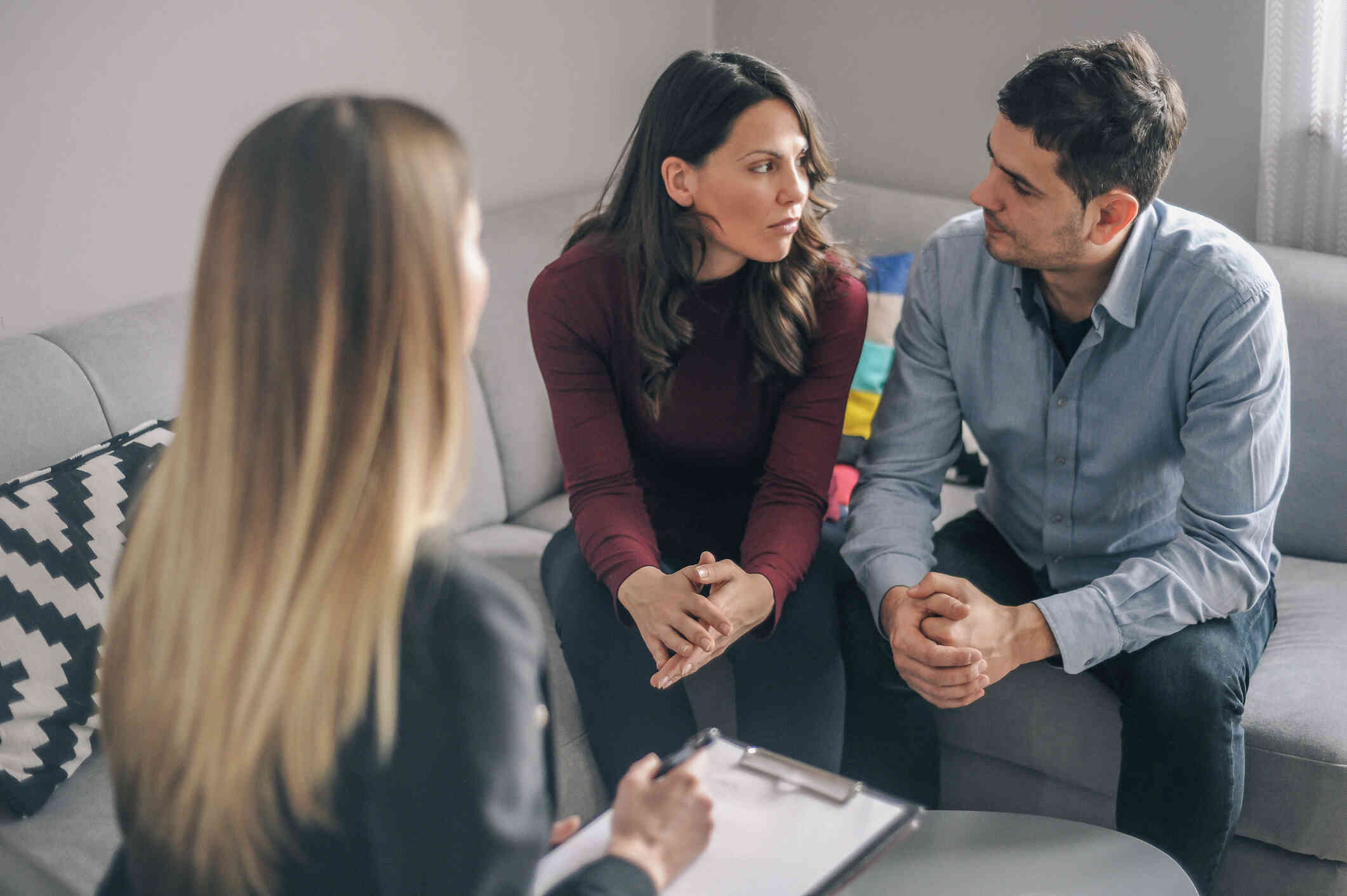 A male and female family memeber sit  next to each other and look at one another with worried expressions as they sit across from their therapist.
